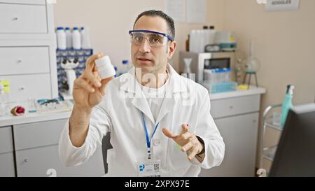 Hispanischer Mann im Labormantel, der eine Flasche im Labor hält, kommuniziert mit einem Kollegen. Stockfoto