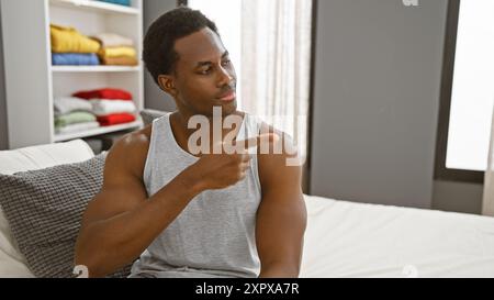 Junger afroamerikanischer Mann, der in einem modernen Schlafzimmer sitzt und auf etwas zeigt Stockfoto