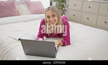 Eine lächelnde junge blonde Frau in Loungewear, die einen Laptop mit Notebook auf einem Bett in einem gemütlichen Schlafzimmer benutzt. Stockfoto