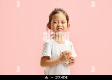 Fröhliches chinesisches Aussehen kleines Mädchen, das ein Glas Milch auf rosa isoliertem Hintergrund hält Stockfoto