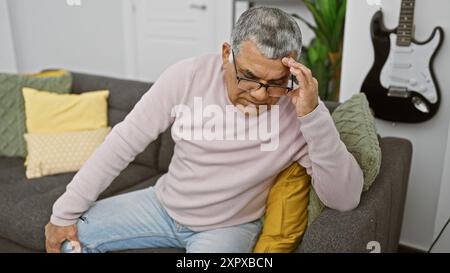 Ein älterer Mann mit grauen Haaren, der eine Brille trägt und sich zu Hause auf einer Couch in einem modernen Wohnzimmer unwohl fühlt. Stockfoto