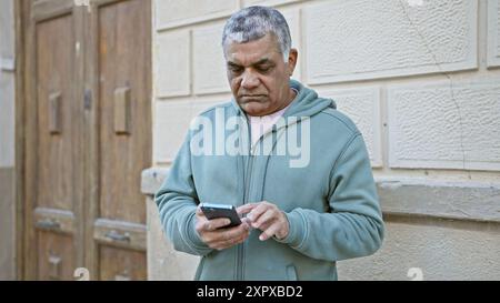 Reifer grauhaariger Mann mit Smartphone auf der Stadtstraße, fokussiert auf den Bildschirm, während er draußen steht. Stockfoto