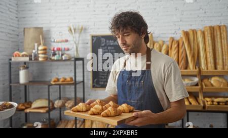 Junger Mann in einer Bäckerei, der ein Tablett mit frischen Croissants hält, umgeben von Regalen mit Brot und Gebäck, während er eine Schürze in einem Innenbereich trägt Stockfoto