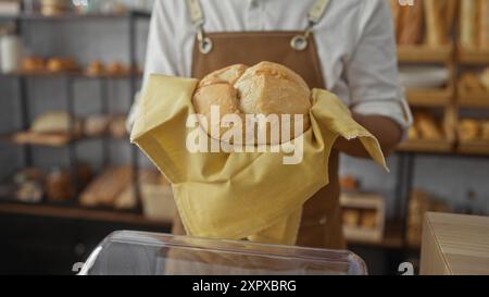 Junger Mann, der frisch gebackenes Brot in einem gelben Tuch in einer Bäckerei mit verschiedenen Brotlaiben im Hintergrund hält Stockfoto