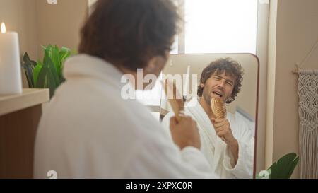 Junger Mann in einer Spa-Umgebung, der in einen Pinsel singt, während er sein Spiegelbild im Spiegel betrachtet, umgeben von Pflanzen und einer zündeten Kerze, die relaxa überträgt Stockfoto