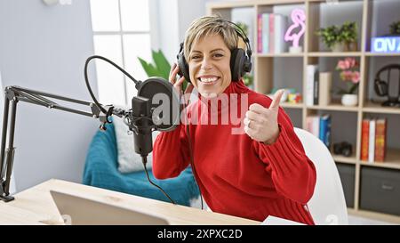 Lächelnde Frau mit kurzen blonden Haaren in einem roten Pullover, der in einem modernen Radio-Studio die Daumen nach oben zeigt Stockfoto