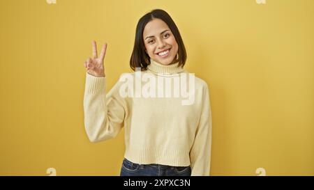 Lächelnde junge hispanische Frau in einem weißen Pullover, die ein Friedenszeichen vor gelbem Hintergrund macht. Stockfoto