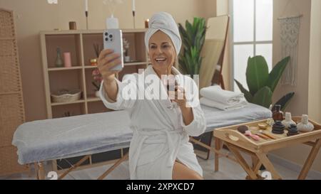 Frau genießt Spa-Sitzung, die Selfie mit Weinglas im Wellnesscenter mit Massagetisch und Kosmetikprodukten nimmt Stockfoto
