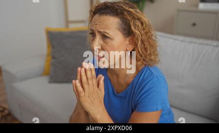 Eine hispanische Frau mittleren Alters mit lockigen Haaren sitzt in einem Wohnzimmer, trägt ein blaues Hemd und wirkt nachdenklich und reflektierend in einem Innenraum. Stockfoto