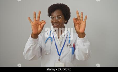 Lächelnde afroamerikanische Ärztin mit lockigen Haaren und Brille, die Zeichen auf beiden Händen auf einem isolierten weißen Hintergrund in Ordnung hält Stockfoto