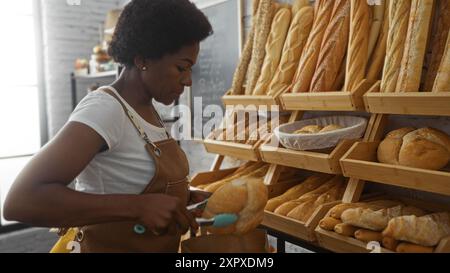 Frau, die in einer Bäckerei arbeitet und frisches Brot in eine Papiertüte legt, umgeben von verschiedenen Brotlaiben auf Holzregalen. Stockfoto