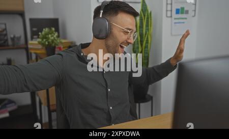 Junger Mann, der im Büro mit Kopfhörern tanzt und dabei Musik genießt und eine unterhaltsame und entspannte Arbeitsumgebung präsentiert Stockfoto