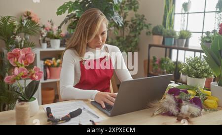 Eine junge Frau arbeitet an einem Laptop in einem pulsierenden Blumenladen, umgeben von grünen Pflanzen und Blumen. Stockfoto