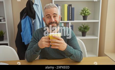 Lächelnder bärtiger Mann mit einem Becher in einem modernen Wohnzimmer mit Bücherregal und Pflanzen. Stockfoto