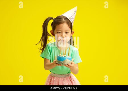 Fokussiertes fünfjähriges chinesisches Aussehen Kind in Party Hut Blow Out Kerzen auf Geburtstagskuchen hält es und steht vor gelbem Hintergrund. Stockfoto