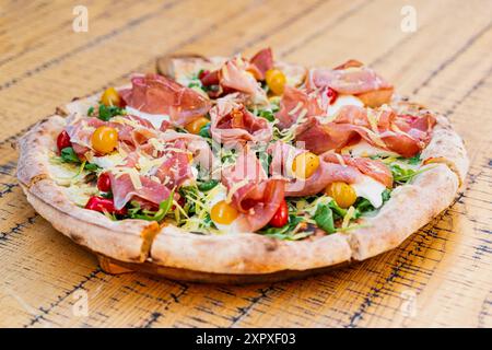 Köstliche handwerkliche Pizza mit dünnen Scheiben Schinken, bunten Kirschtomaten und frischem Rucola. Serviert auf einem rustikalen Holztisch, perfekt Stockfoto