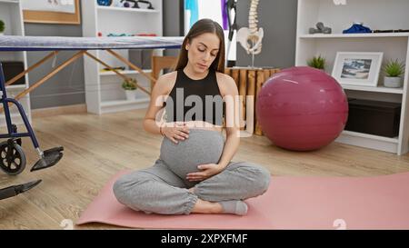 Eine schwangere hispanische Frau in Yogakleidung, die in einem Rehabilitationszentrum mit physiotherapeutischen Werkzeugen meditiert. Stockfoto