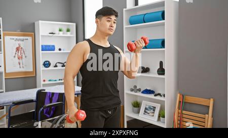 Ein junger asiatischer Mann übt mit Kurzhanteln in einer modernen Rehabilitationsklinik. Stockfoto