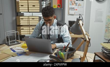 Ein konzentrierter junger Mann, der an einem Laptop in einem überfüllten Detektivbüro arbeitet, voll mit Akten und Beweisen. Stockfoto