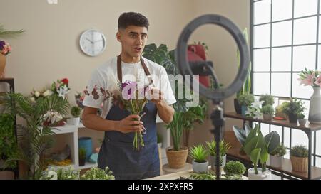 Ein junger Mann mit Schürze arrangiert Blumen in einem lichtdurchfluteten Blumenladen, umgeben von verschiedenen Pflanzen und einem Ringlicht. Stockfoto