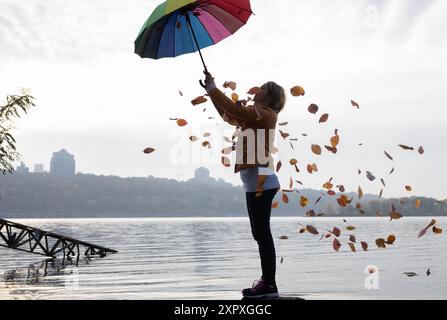 An einem bewölkten Tag hat eine Frau Spaß, unter einem Regenbogenschirm zu stehen, aus dem herbstliche Blätter fallen. Herbstatmosphäre. Saisonale Freuden und Spaziergänge. H Stockfoto