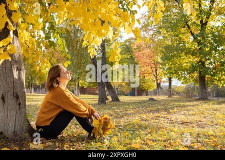 Die 35-40-jährige Frau in einem orangefarbenen Pullover an einem hellen, warmen, sonnigen Tag sitzt träumerisch auf dem Boden und genießt den Moment. Inspiration, Energie der Natur. Stockfoto