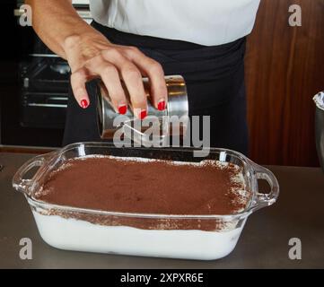 Eine Person, die Kakaopulver über einem hausgemachten Tiramisu-Dessert in einer Glasschale bestäubt, mit einer Rührschüssel an der Seite. Stockfoto