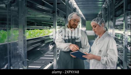 Ein paar Landwirtschaftsexperten, die bei Indoor Vertical Farm arbeiten. Junges Team von Gartenspezialisten für Schutzkleidung und Kopfbedeckungen, die in einem Flur mit frischen Produkten sprechen Stockfoto
