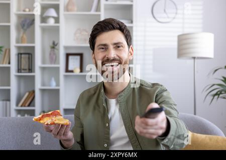 Fröhlicher Mann, der ein Stück Pizza hält, während er zu Hause fernsieht. Lächelnder Erwachsener, der sich auf dem Sofa mit Fernbedienung und leckerem Snack entspannt und die Freizeit genießt Stockfoto