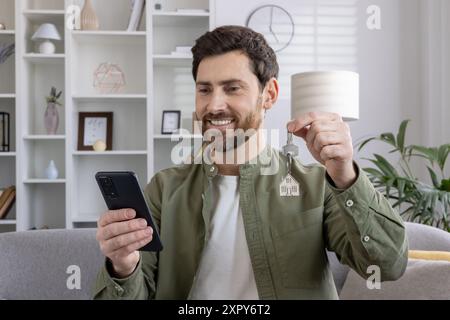 Lächelnder Mann, der neue Hausschlüssel hält, während er auf das Telefon blickt. Zeigt Aufregung, Immobilienerfolg, neues Wohneigentum und modernen Lebensstil. Leistungskonzept und digitale Kommunikation. Stockfoto