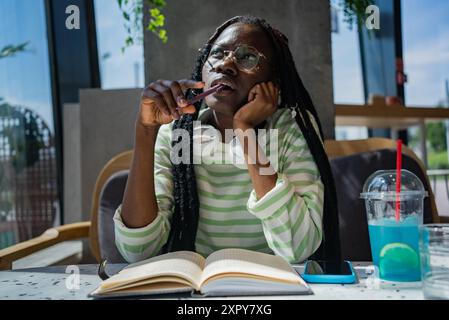 Nachdenkliche junge afroamerikanische Frau mit Zöpfen und Brillen, die in einem Café in einem Notizbuch schreibt, mit Kopfhörern um den Hals und einem Drink in der Nähe. Stockfoto