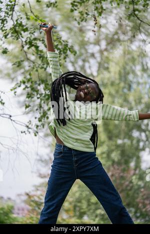 Glückliche junge afroamerikanische Frau mit langen Zöpfen und Brillen, die fröhlich hüpft, während sie ein Smartphone in einem üppigen grünen Park hält. Stockfoto
