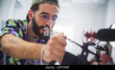 Fokussierter junger männlicher Kameramann mit Bart und Brille passt die Kamera während einer Innenaufnahme an und hält die Essenz moderner Filmkunst und kreativer Werbung in Einem professionellen Setting fest. Stockfoto