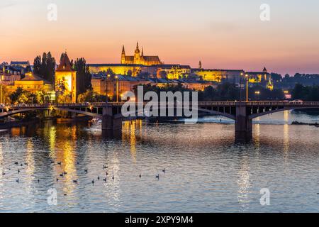 Ein Blick auf die Prager Burg, beleuchtet in der Abenddämmerung, von einer Brücke über die Moldau aus gesehen. Die Lichter der Stadt reflektieren das ruhige Wasser und schaffen eine ruhige und malerische Szene. Prag, Tschechien Stockfoto