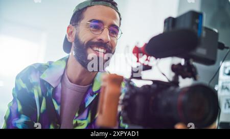 Smiling Young Latin Male Filmemacher, der Eine professionelle Kamera in Einem gut beleuchteten Studio betreibt und ansprechende Inhalte für Ein modernes Werbe- oder Filmproduktionsprojekt aufnimmt. Stockfoto