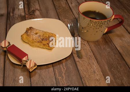Blätterteig gefüllt mit Äpfeln auf einem weißen Teller mit einer Tasse Kaffee und einem kleinen Geschenk am Rand des Tellers Stockfoto