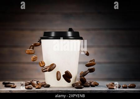 Kaffee in einer Papiertasse steht auf einem Holztisch, geröstete Kaffeebohnen fliegen um den Papierbecher. Kaffeebohnen auf einem Holztisch. Heißer Kaffee im Papier, Stockfoto