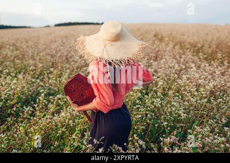 Rückansicht einer stilvollen Frau mit Strohhut auf dem blühenden Sandweatfeld bei Sonnenuntergang, die Lederhandtasche hält. Modische Kleidung und Accessoires für den Sommer Stockfoto