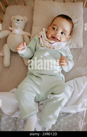 Kurioses männliches Baby, das auf dem Rücken im Babybett liegt und seine Mutter ansieht, während es weit lächelt Stockfoto