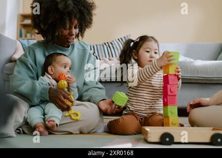 Afroamerikaner-Mutter hält ihren kleinen Sohn fest, während sie asiatischen Kleinkindern hilft, einen Backsteinturm zu bauen Stockfoto