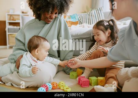 Junge, der die Hände eines asiatischen Mädchens und ihrer Mutter berührt, während seine afroamerikanische Mutter sie beobachtet und lächelt Stockfoto