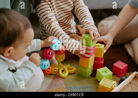 Aus einem hohen Winkel gezogene Kinderhände, die bunte Ziegelsteine halten und rasseln, während die Erwachsene Hand ihnen beim Bau des Turms hilft Stockfoto