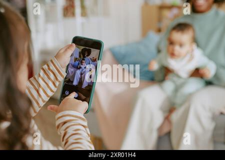 Mittlere Nahaufnahme von kleinen Händen eines Mädchens, das Telefon hält, um ein Foto von Baby mit seiner afroamerikanischen Mutter zu machen, während sie auf dem Sofa sitzen Stockfoto