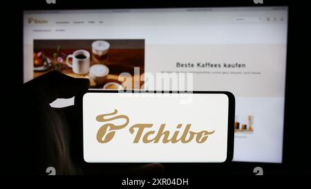 Person, die ein Mobiltelefon mit dem Logo der deutschen Kaffeehändler Tchibo GmbH vor der Business-Website hält. Schwerpunkt auf dem Display des Telefons. Stockfoto