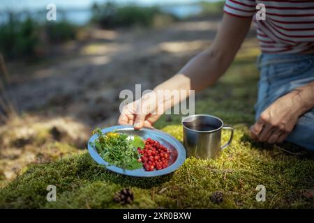 Unbekannte Frau in der Natur mit Campingbecher, Teller mit Beeren und Minze Braut leckeren Tee. Stockfoto