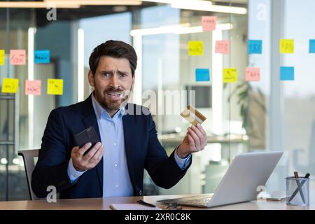 Geschäftsmann im Büro mit Kreditkarte und Telefon, der frustriert aussieht. Mann mit Anzug, der am Schreibtisch sitzt, mit Laptop und Haftnotizen auf Glas im Hintergrund. Stockfoto