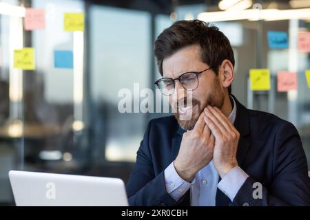 Geschäftsmann im Büro mit Zahnschmerzen während der Arbeit am Laptop. Mann mit Wange bei Schmerzen und Unwohlsein, was auf Zahnprobleme während der Arbeitszeit hinweist. Stockfoto