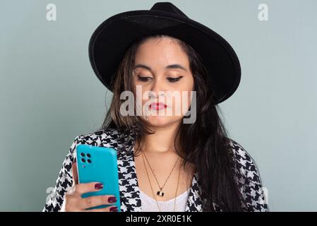 Junge schöne Frau, die im Studio steht und auf das Handy schaut. Trägt schwarzen Hut vor hellgrünem Hintergrund. Stockfoto