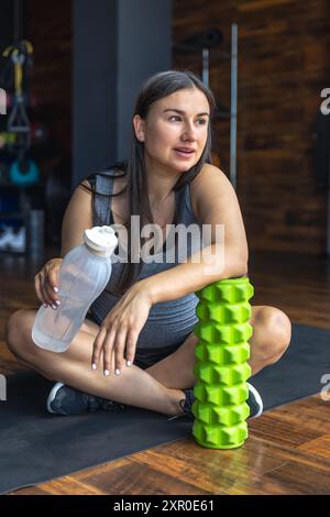 Eine schwangere Frau im Fitnessstudio sitzt auf einer Matte und trinkt Wasser. Stockfoto