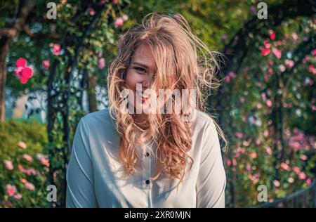 Eine junge Frau mit lockigem Haar genießt einen sonnigen Tag in einem wunderschönen Garten voller blühender Rosen Stockfoto